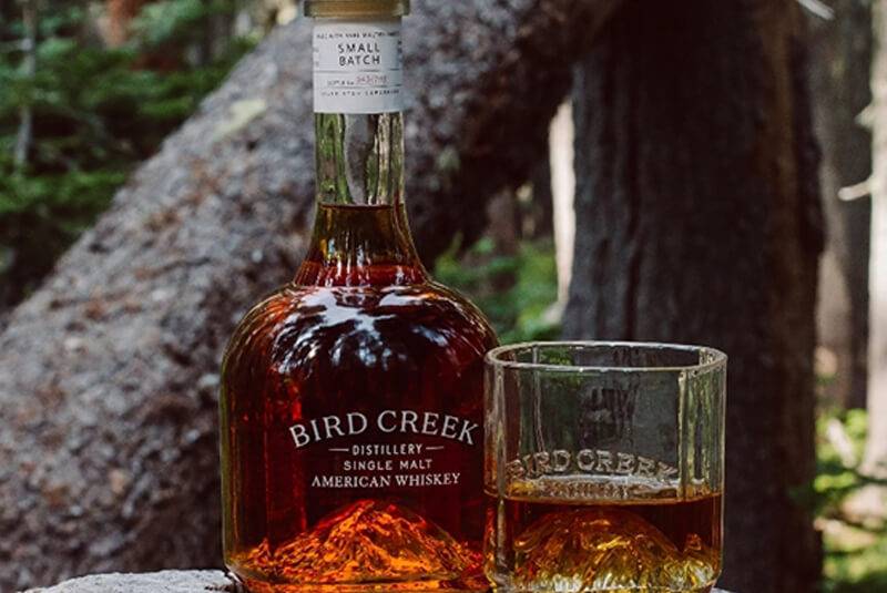 Bird Creek single malt whiskey bottle and glass on a rock in a forest